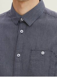 Camicia button-up di colore blu navy con un motivo testurizzato e a piccole righe. Presenta un colletto, bottoni bianchi e una tasca sul lato sinistro del petto.