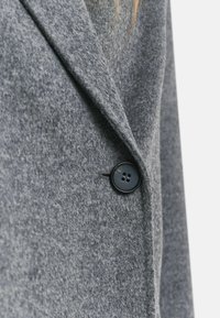 Grijze wollen jas met een gestructureerde kraag, voorzien van een zwarte sluiting met vier knopen. Dikke, zachte textuur met een subtiel patroon.
