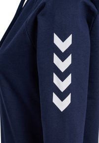 Mörkblå sweatshirt i strukturerad tyg. Har en vit zigzag-grafik längs vänster ärm och en huva med dragsko.