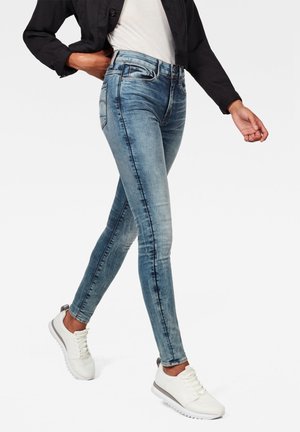 Personne marchant portant un jean skinny bleu délavé, des baskets blanches, une chemise blanche et une veste noire devant un fond clair uni.