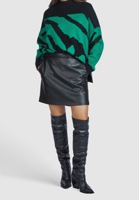 Kobieta ubrana w sweter w zielono-czarny abstrakcyjny wzór, czarną skórzaną spódniczkę mini oraz czarne wysokie botki na obcasie, stojąca z rękami w kieszeniach.