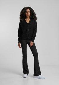 Zwarte gebreide cardigan met een V-hals en vijf knopen, gecombineerd met zwarte flared broek en witte sneakers. Soepele textuur en ontspannen pasvorm.