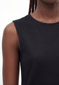Close-up of a person wearing a black sleeveless top with braided hair visible against a white background.