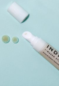 Tubetto di crema per gli occhi bianco con una punta di erogazione arrotondata, accompagnato da due piccole gocce traslucide su uno sfondo azzurro chiaro.