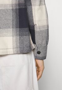 Bawełniana koszula w kratę w odcieniach granatu, szarości i bieli, z podwiniętym mankietem i czarnym guzikiem na rękawie.