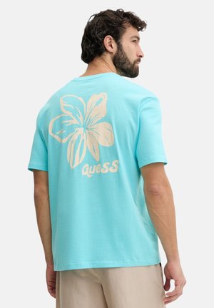 Uomo che indossa una maglietta azzurro chiaro con un grande fiore beige e il logo "Guess" sul retro, abbinata a pantaloni beige, visto di profilo.