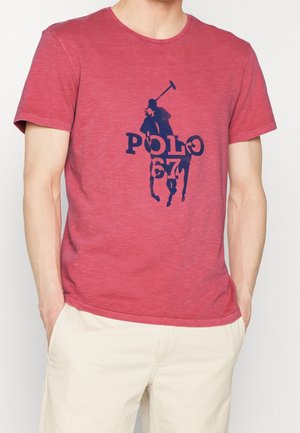 Mężczyzna w czerwonym T-shircie z krótkim rękawem, z granatowym nadrukiem gracza polo i napisem "POLO 67", w połączeniu z jasnobeżowymi szortami.