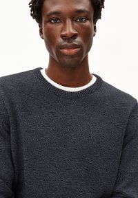 Pull-over en tricot bleu marine avec une surface texturée, col rond côtelé, et une chemise blanche superposée en dessous, présentant un design décontracté.