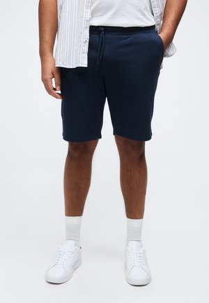 Άντρας που φοράει σκούρα μπλε σορτς με κορδόνι, λευκά αθλητικά παπούτσια με λευκές κάλτσες, λευκό T-shirt και ριγέ ανοιχτό πουκάμισο, όρθιος με τα χέρια στις τσέπες.