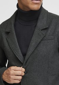 Blazer de lana gris con solapa, cierre de un solo botón y un bolsillo en el pecho, superpuesto sobre un cuello de tortuga negro de canalé.