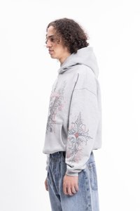 Sivá mikina s kapucňou a grafickým dizajnom, ktorý zdobí zložitý vzor na rukáve. Predná kapsa a voľný strih. Nosené s svetlomodrými džínsami.