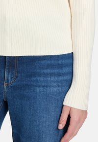 Gerippter cremefarbener Pullover mit langen Ärmeln, getragen über blauen Jeans. Der Pullover hat einen geraden Saum und strukturierte Stoff.