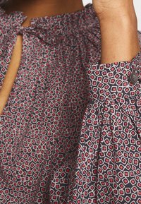 Blouse en tissu à motif présentant un design géométrique en bleu marine, rouge et blanc. Comprend un col avec cordon de serrage et des poignets boutonnés.