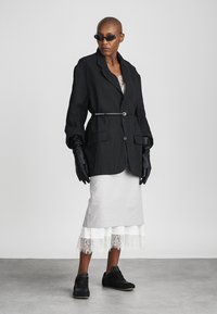 Osoba nosi oversized crni blazer s tankim pojasom, crne kožne rukavice, suknju od bijelog čipkastog ruba, crne tenisice i uske crne sunčane naočale.