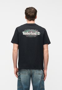 Άνδρας που φοράει μαύρο μπλουζάκι Timberland με πράσινο και λευκό λογότυπο στο πίσω μέρος, συνδυασμένο με μπλε τζιν παντελόνι cargo, όρθιος με την πλάτη γυρισμένη.