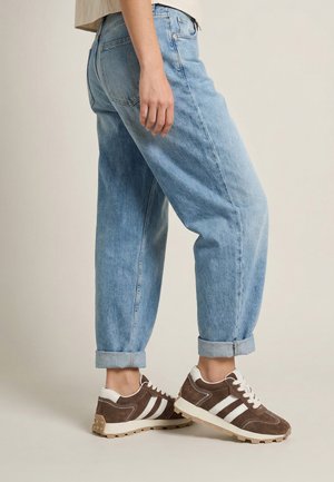 Persoon draagt lichtblauwe opgerolde spijkerbroek en bruine en witte veter sneakers, staand tegen een effen beige achtergrond.