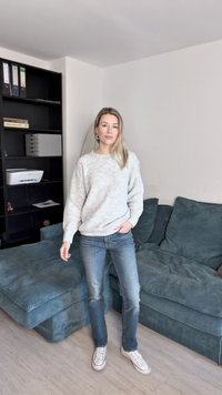 Lichtgrijze, gestructureerde sweater met een ronde hals, gecombineerd met een blauwe spijkerbroek en witte sneakers, staande in een gezellige woonkamer.