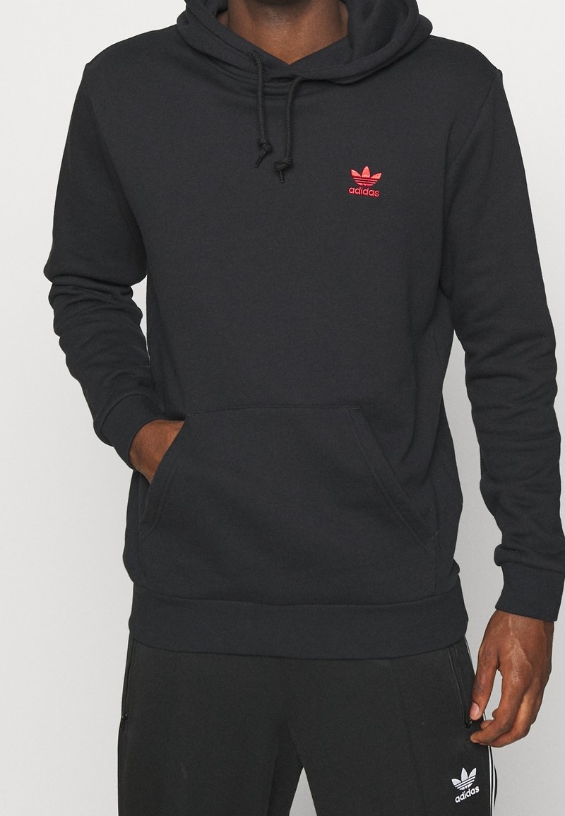 Svart huvtröja i mjukt tyg med en framficka, dragsko i huvan och liten röd Adidas-logotyp på bröstet.