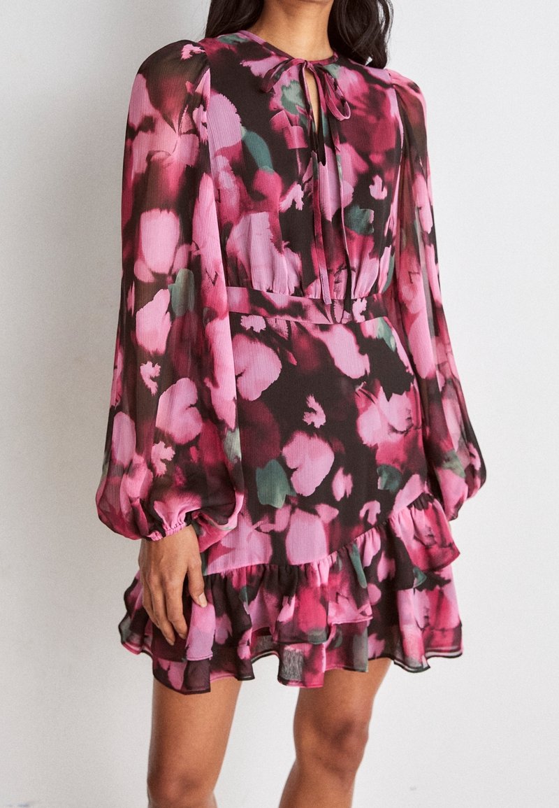 Femme portant une robe longue à manches longues rose et noire à motifs floraux, avec un ourlet à volants et un petit col en forme de goutte noué avec un nœud.