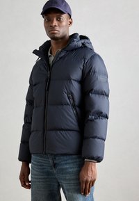 Navy puffer jacket with hood, zipper closure, and prominent stitching. Worn over a gray shirt, paired with blue jeans.
