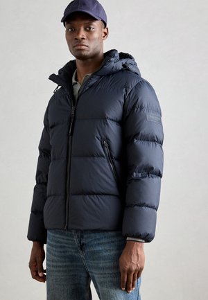 Marineblauwe pufferjack met capuchon, ritssluiting en opvallende steeknaden. Draagt over een grijs shirt, gecombineerd met blauwe spijkerbroek.