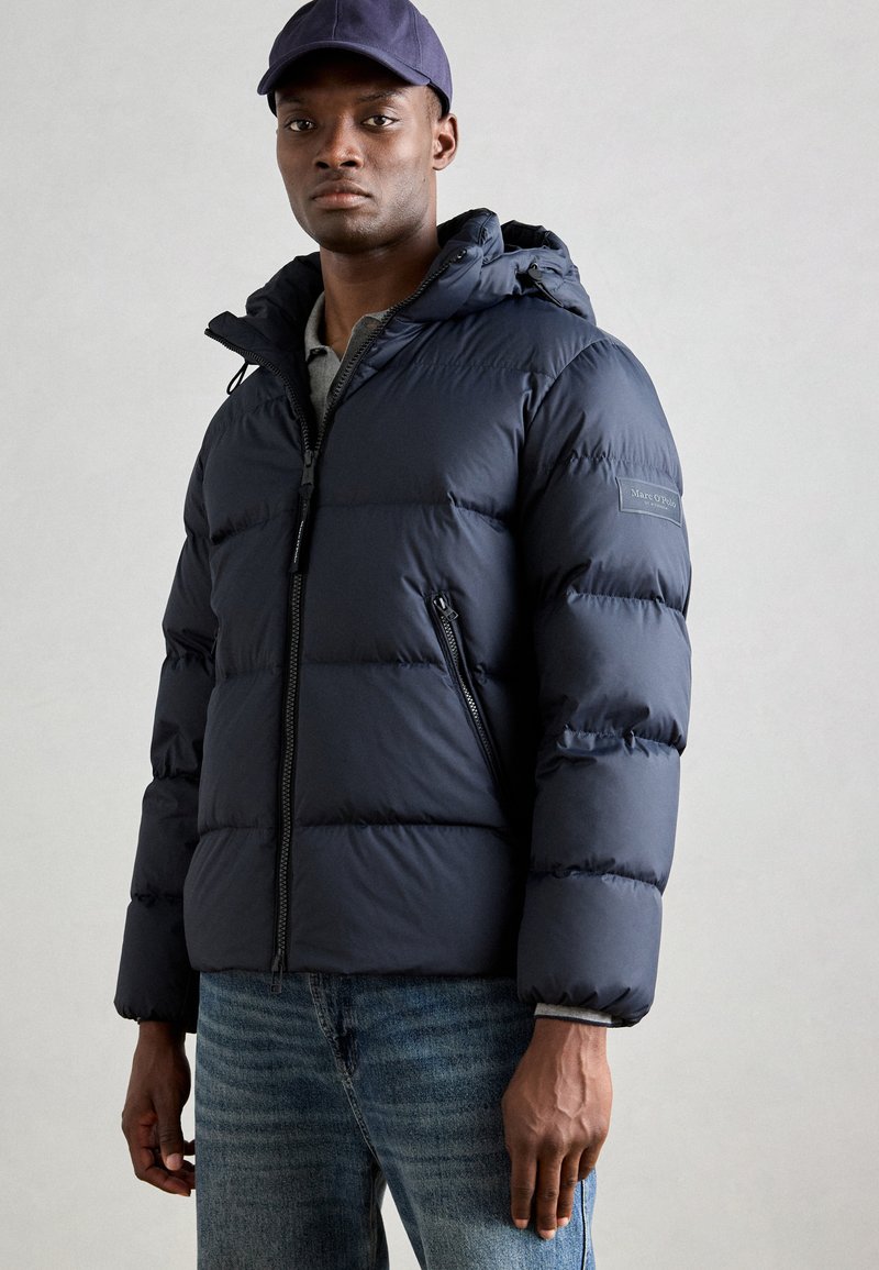 Navy puffer jacket with hood, zipper closure, and prominent stitching. Worn over a gray shirt, paired with blue jeans.