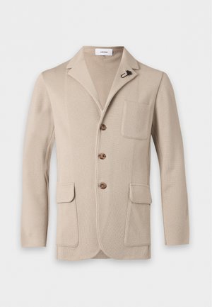 Jeune homme portant un blazer beige par-dessus une chemise à carreaux, un pantalon noir et des chaussures noires, debout avec une main dans la poche sur fond blanc.