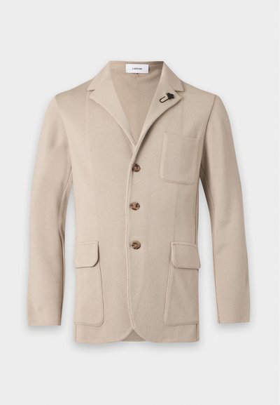 Blazer en maille beige avec revers crantés, trois boutons marron, une poche poitrine, deux poches avant à rabat et une épingle florale noire sur le revers.
