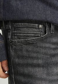 Črne denim hlače z teksturiranim zaključkom, ki vsebujejo pet žepov in diskretno šivanje. Na zadnjem žepu je vidna kovinska oznaka.