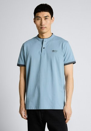 Mężczyzna w jasnoniebieskiej koszulce typu henley z krótkim rękawem i napisem "19/62" na piersi, stojący na tle jednolitego jasnoszarego tła.