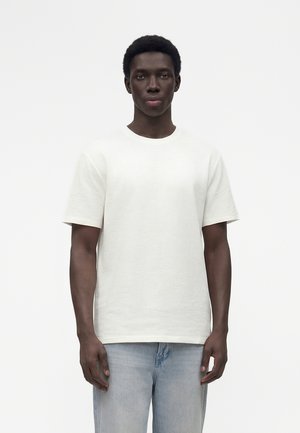 Joven con expresión neutra, vestido con una camisa blanca texturizada de manga corta y vaqueros azul claro, de pie frente a un fondo liso.