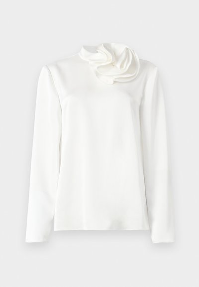 Blusa bianca a maniche lunghe con collo alto e un grande dettaglio floreale in tessuto sulla spalla sinistra, su uno sfondo semplice.