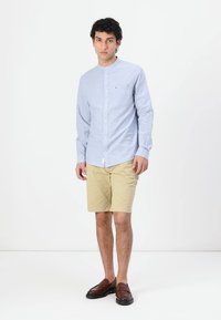 Camicia a maniche lunghe azzurro chiaro con colletto, abbinata a pantaloni beige e scarpe slip-on marroni. Presenta un piccolo logo sulla camicia.