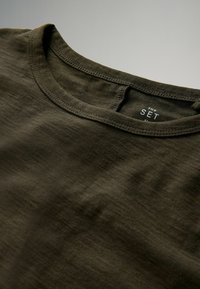 Olivegrünes T-Shirt mit Rundhalsausschnitt aus weichem Stoff, das eine glatte Textur mit subtilen Variationen aufweist und an einem Etikett mit der Aufschrift "THE SET" versehen ist.
