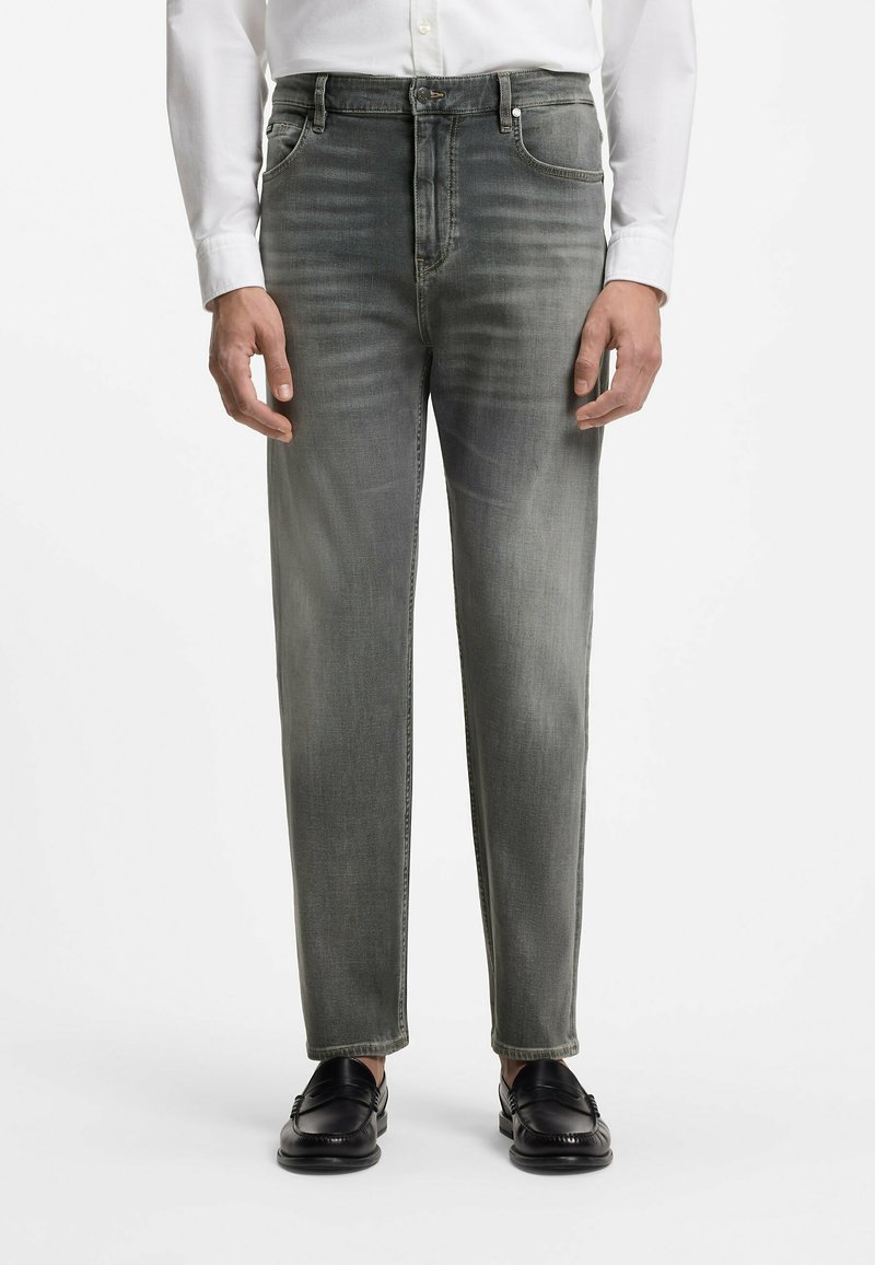 Homme portant un jean gris délavé, une chemise blanche et des mocassins noirs, debout devant un fond blanc.