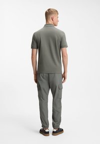 Homme aux cheveux blonds courts portant un polo gris à manches courtes, un pantalon cargo gris et des baskets noires, vu de dos sur fond blanc.