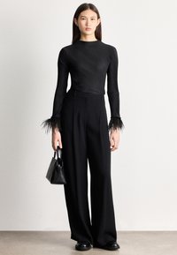 Žena oblečená v čiernom rebrovanom topu s perovými manžetami, široké čierne nohavice, čierne topánky a drží malú čiernu kabelku.