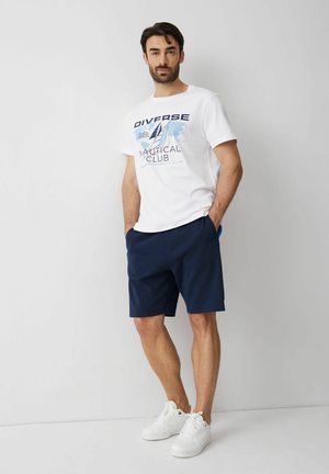 Mężczyzna stoi oparty o ścianę, ubrany w biały graficzny T-shirt w stylu morskim, granatowe szorty i białe trampki, ręce w kieszeniach, zrelaksowana poza.