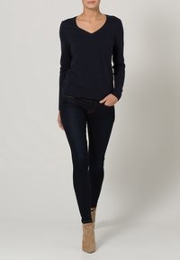 Marineblaue Langarm-V-Ausschnitt-Bluse kombiniert mit dunklen Skinny-Jeans, ergänzt durch braune Knöchelstiefel. Glatter Stoff mit figurbetontem Schnitt.