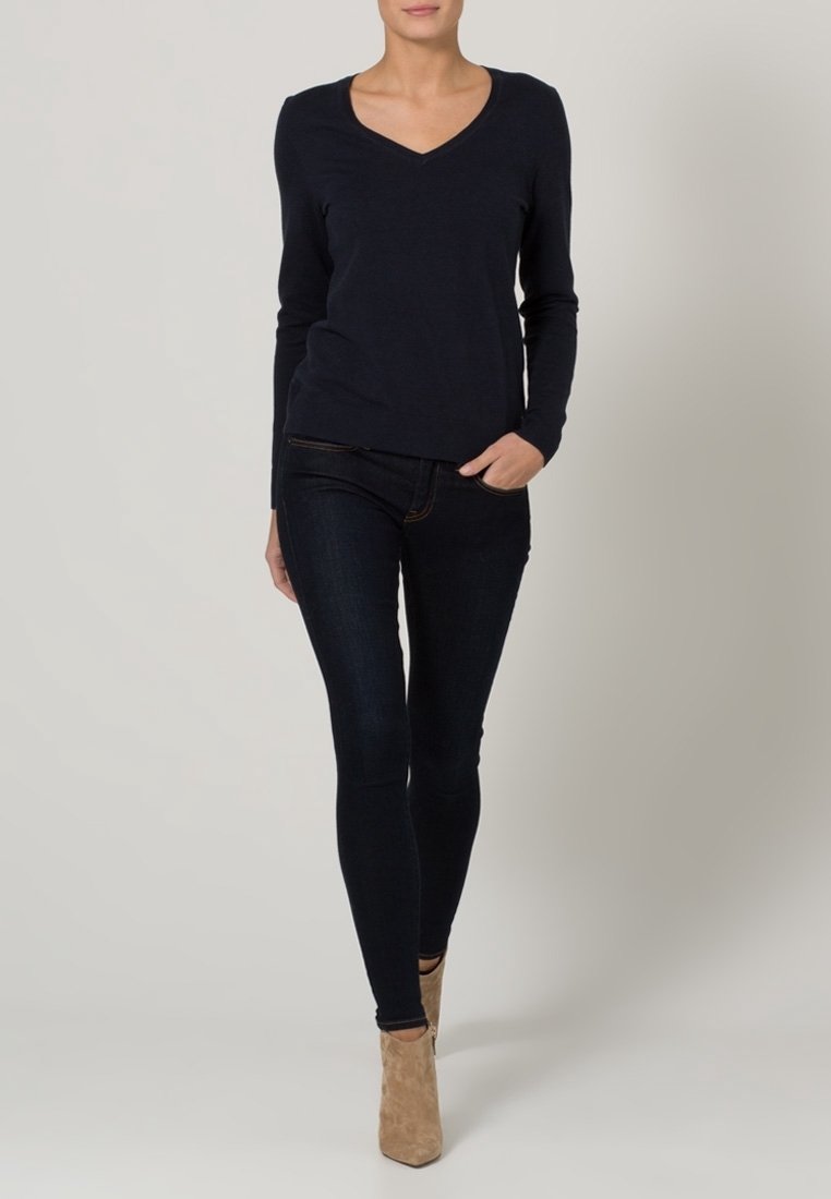 Marineblaue Langarm-V-Ausschnitt-Bluse kombiniert mit dunklen Skinny-Jeans, ergänzt durch braune Knöchelstiefel. Glatter Stoff mit figurbetontem Schnitt.