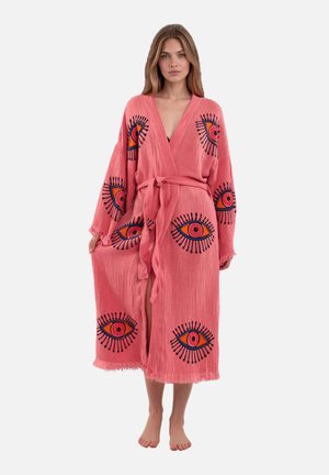 Vrouw draagt een roze gestructureerde badjas met grote kleurrijke oogmotieven, met ceintuur in de taille, staat op blote voeten voor een effen achtergrond.