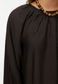 Blusa nera di tessuto testurizzato con maniche lunghe e scollatura arricciata. Accessoriata con una collana di perline di legno rotonde in toni naturali.