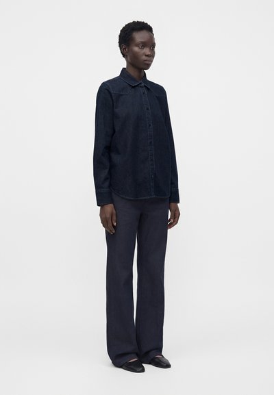 Femme debout portant une chemise à manches longues bleu foncé boutonnée, un pantalon ample assorti et des chaussures noires sur un fond clair uni.