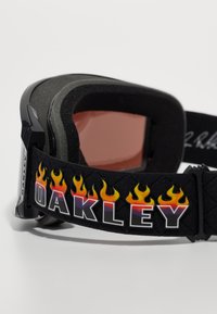 Juodi slidinėjimo akiniai su putų interjeru, raudonais objektyvais ir reguliuojama juosta, kurioje yra ugnies grafika ir tekstas "Oakley".