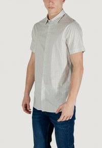 Camicia a maniche corte con bottoni di colore grigio chiaro, con un motivo geometrico, dotata di un colletto strutturato e una texture di tessuto liscia, abbinata a jeans blu.