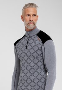 Grå langærmet top med høj krave, præget af et diagonalt mønster med ordet "Dovre." Sorte accenter på skuldrene. Fleksibelt stof.