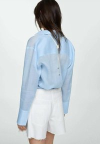 Chemise button-up légère bleu clair, transparente, avec des manches longues et des poignets contrastants, associée à un short blanc avec des bords effilochés.