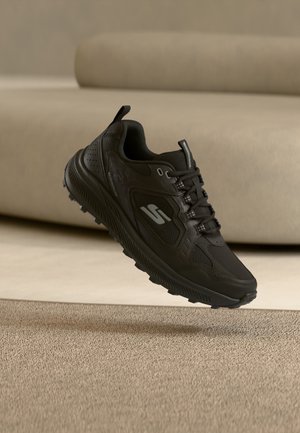 Schwarzer Sportschuh mit strukturierter Sohle und Schnürsenkeln, mit silbernem Logo, schwebt über einem teppichbedeckten Boden nahe einem beigen Sofa.