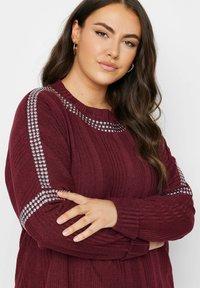 Burgundy stickad tröja med unik ribbad textur och silverfärgade nitar längs axlarna. Rund halsringning och långa ärmar.