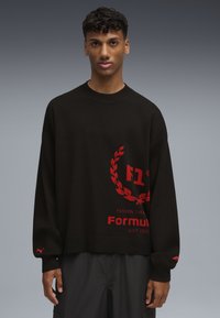 Felpa nera a maglia con un logo circolare rosso con design di alloro, testo "F1" e scritta "PASSION THEME" sul davanti.
